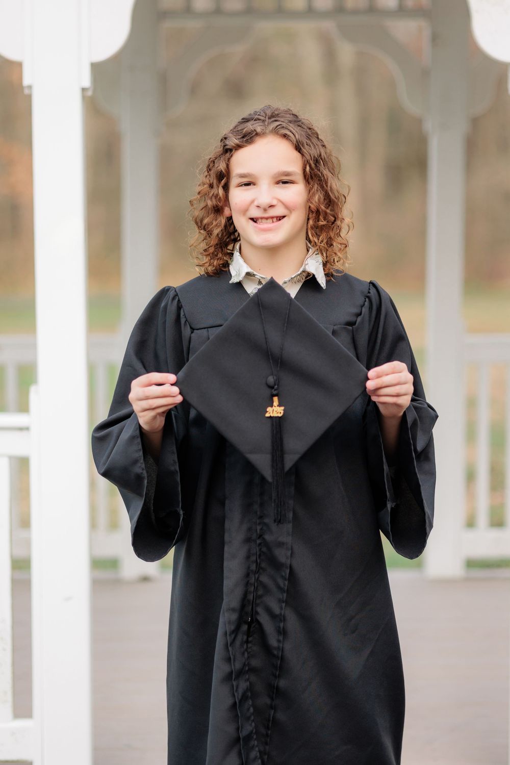 Smiling graduate in black gown holding mortarboard cap outdoors near white columns.