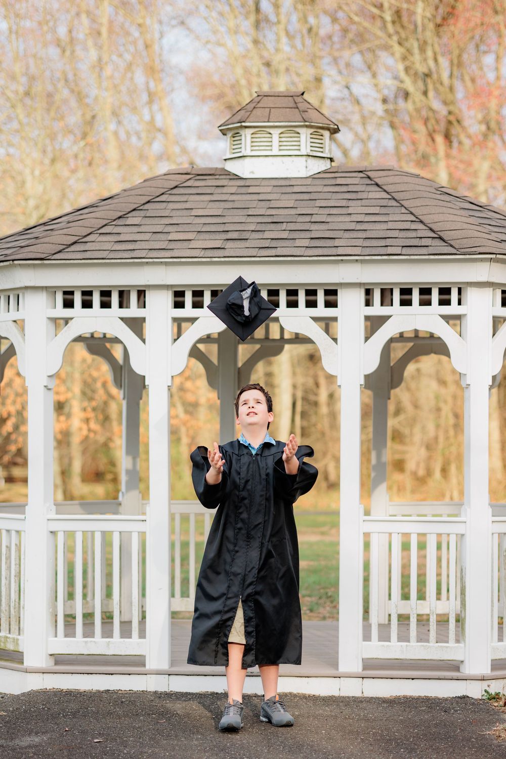 Young graduate in black gown tossing cap in air before white gazebo with autumn trees backdrop.