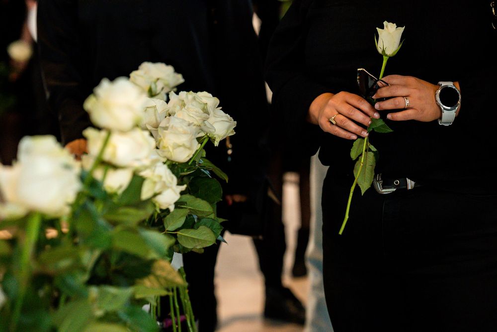 Person in black attire holding a single white rose at a formal or funeral ceremony with floral arrangements.