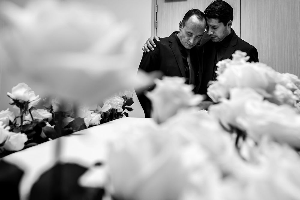 Two men embrace in grief surrounded by white flowers at a funeral service in black and white photo.