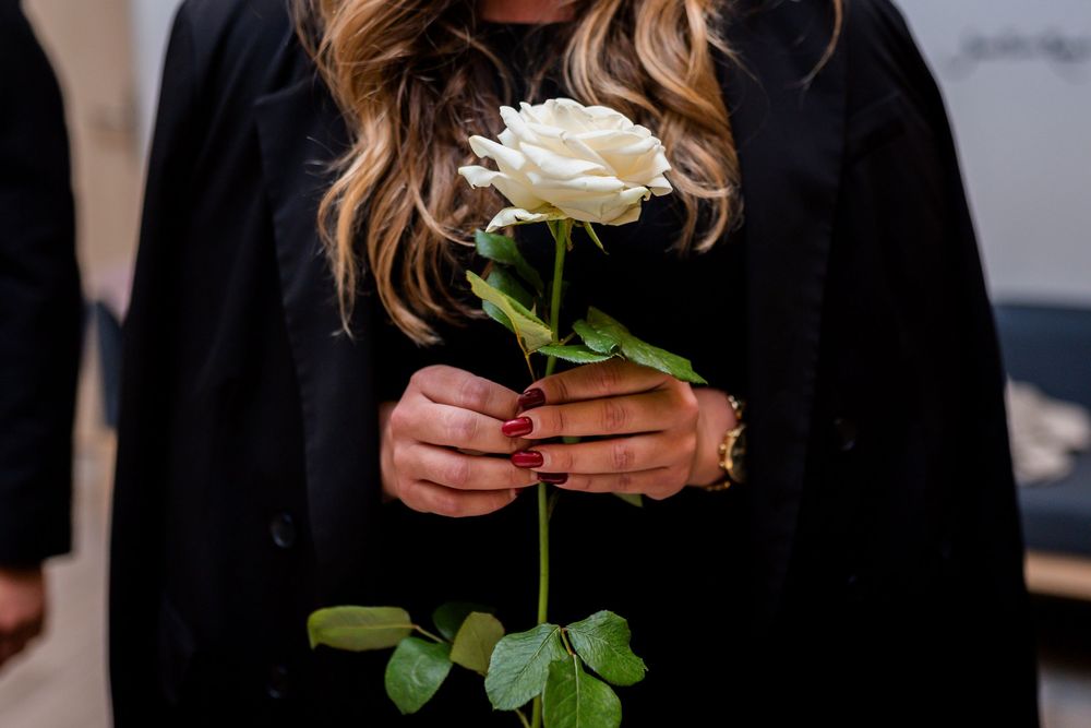 Woman in black outfit holding a single white rose with red manicured nails and gold bracelet.