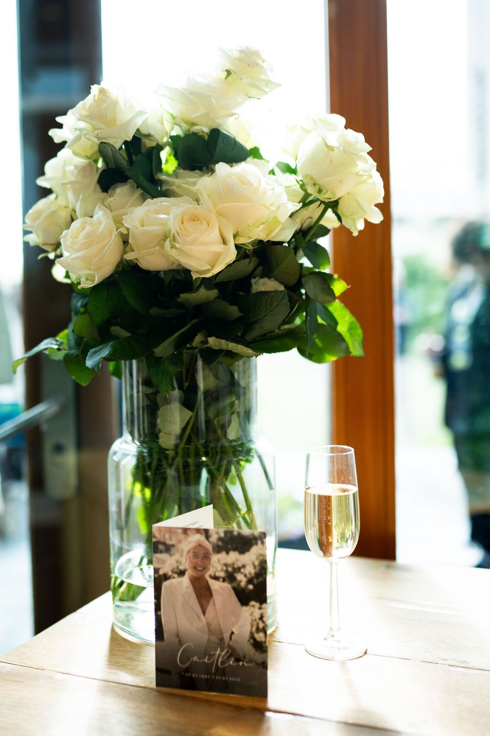 White roses in glass vase beside champagne flute and greeting card on wooden table near window.