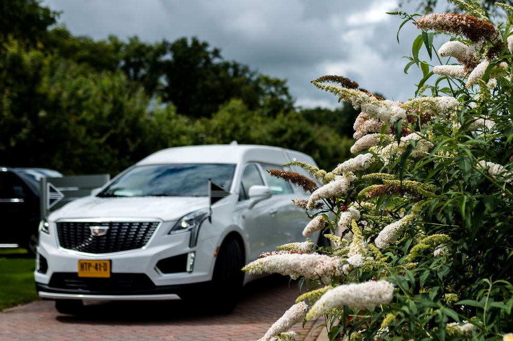 White Cadillac XT5 hearse parked near blooming white butterfly bush under cloudy sky.