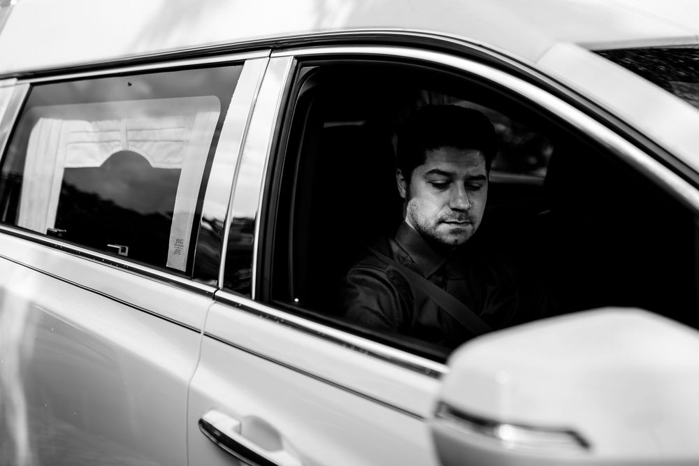Black and white photo of a man sitting in the driver seat of a white car, looking downward.