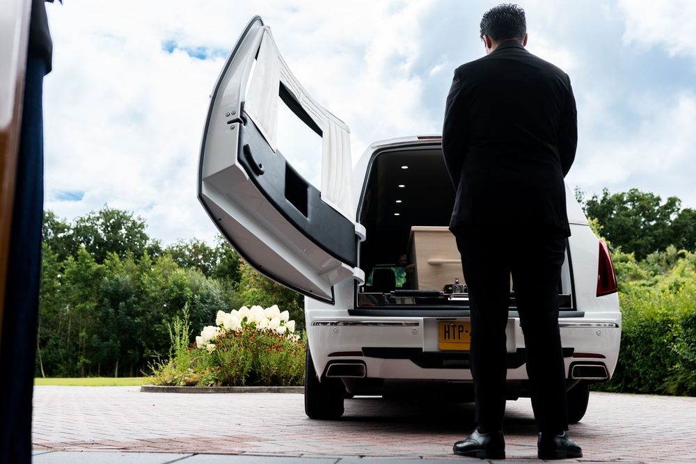Funeral director standing beside an open hearse with a casket at a serene outdoor venue.