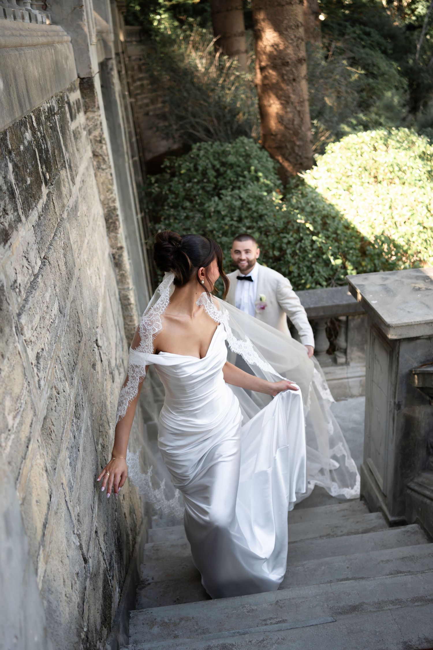 Bride in white gown ascending stone stairs with groom in tuxedo following behind at an elegant outdoor venue.