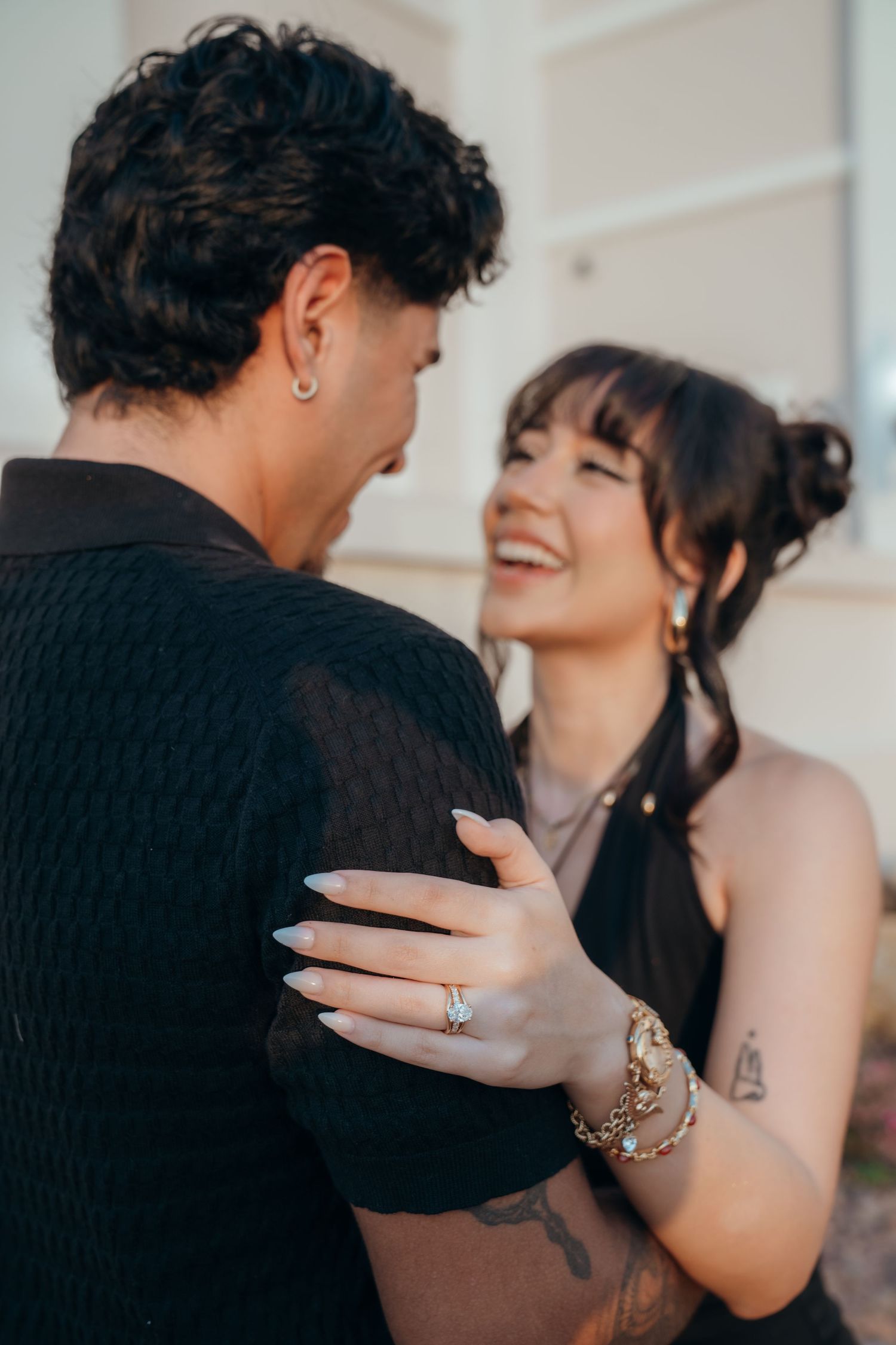 Smiling couple in black outfits sharing a romantic moment, woman wearing ring and bracelet with tattoos visible.