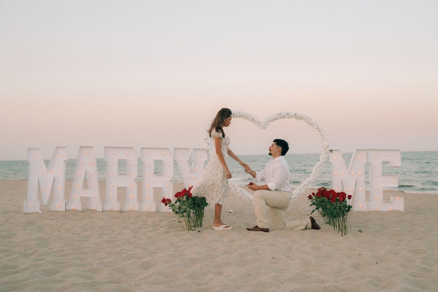 Man proposes to woman on beach with large MARRY ME letters and heart arch decorated with roses at sunset.