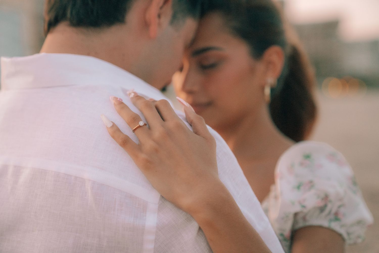 Couple embracing closely, woman wearing diamond engagement ring, soft romantic bokeh background.