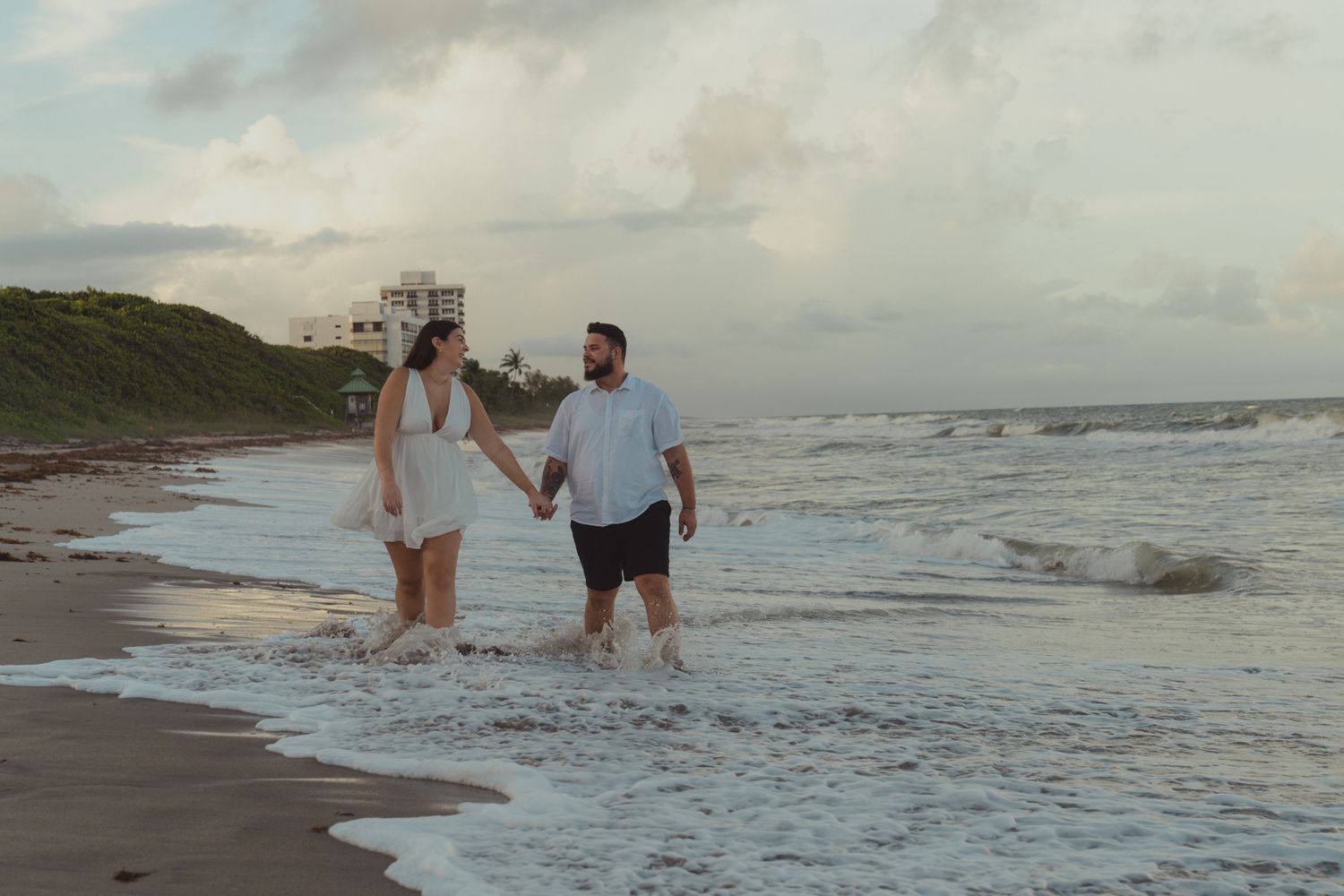 Couple holding hands walking along a sandy beach shore at sunset with ocean waves and cloudy sky.