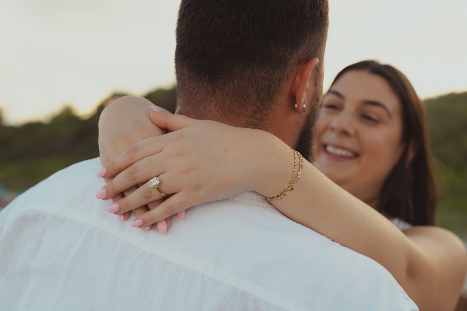 Smiling woman with engagement ring embracing man outdoors at sunset.