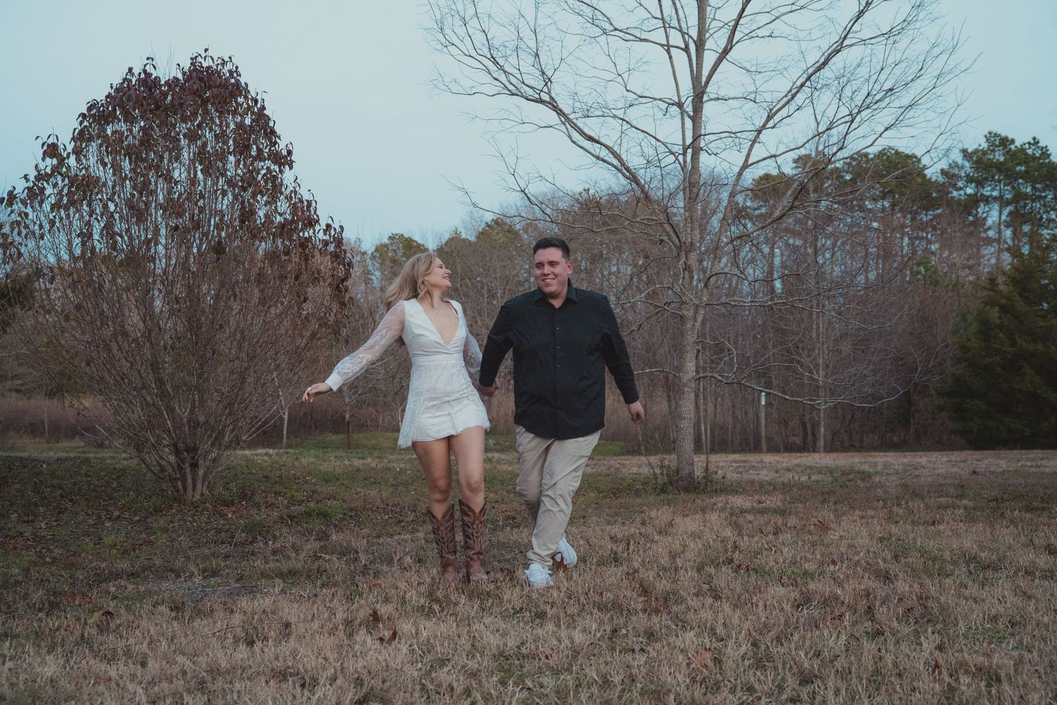 Couple walking hand-in-hand through a rustic field with bare trees, woman in white dress and boots, man in black shirt.