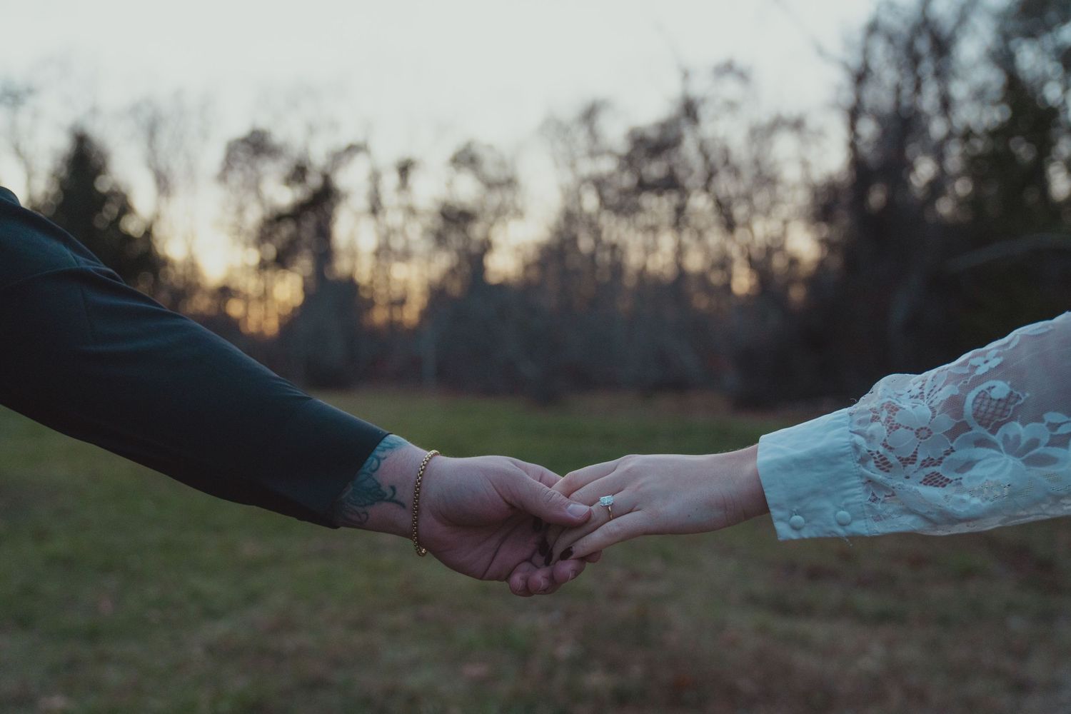 Couple holding hands at sunset in a field, man with tattoo and woman in lace dress, romantic outdoor scene.