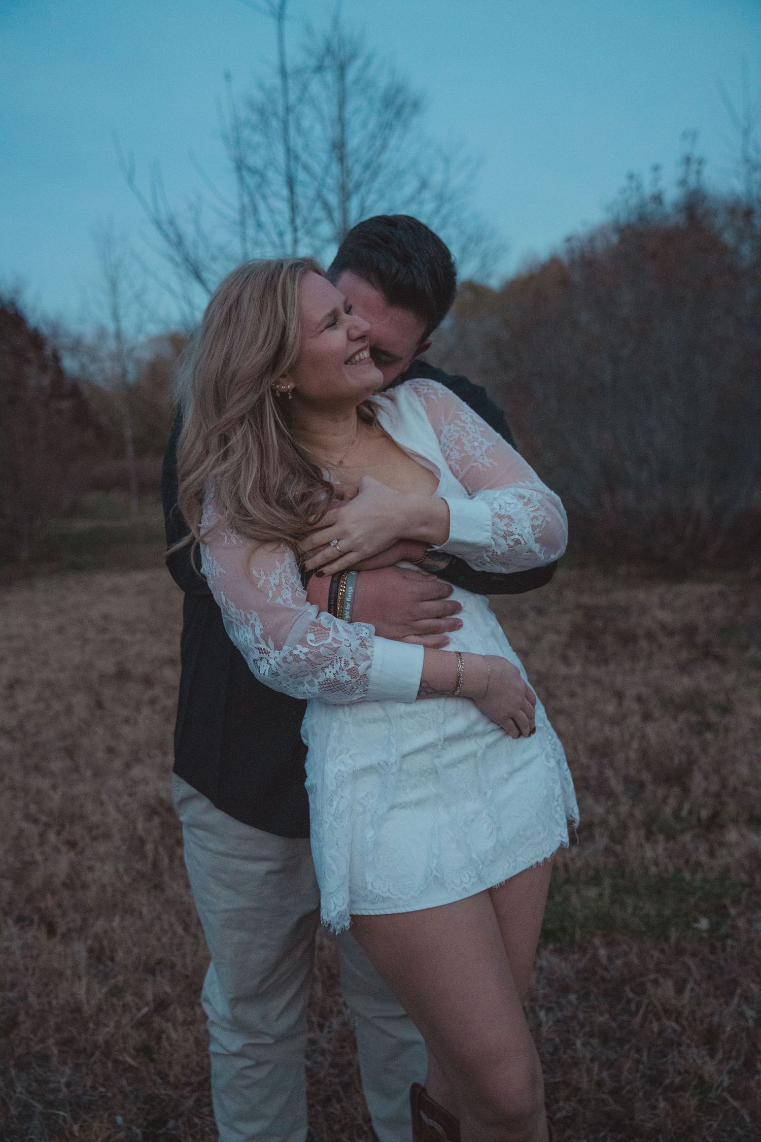 Couple embracing outdoors at dusk, woman in white lace dress smiling as man holds her close in a romantic field setting.
