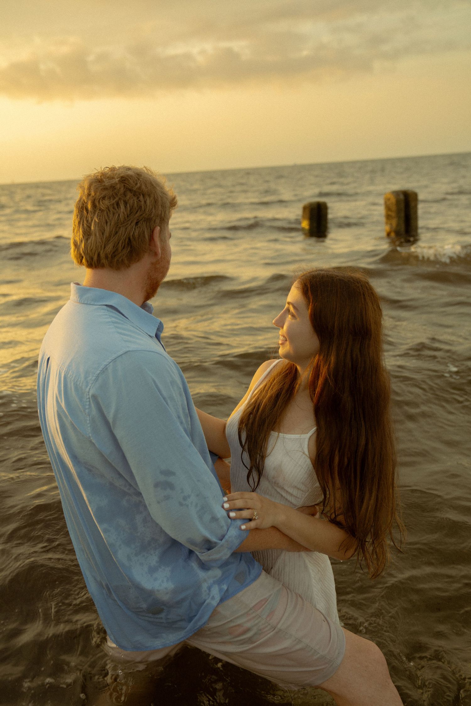 Couple embracing in ocean water at golden sunset, woman in white dress gazing up at man in blue shirt.