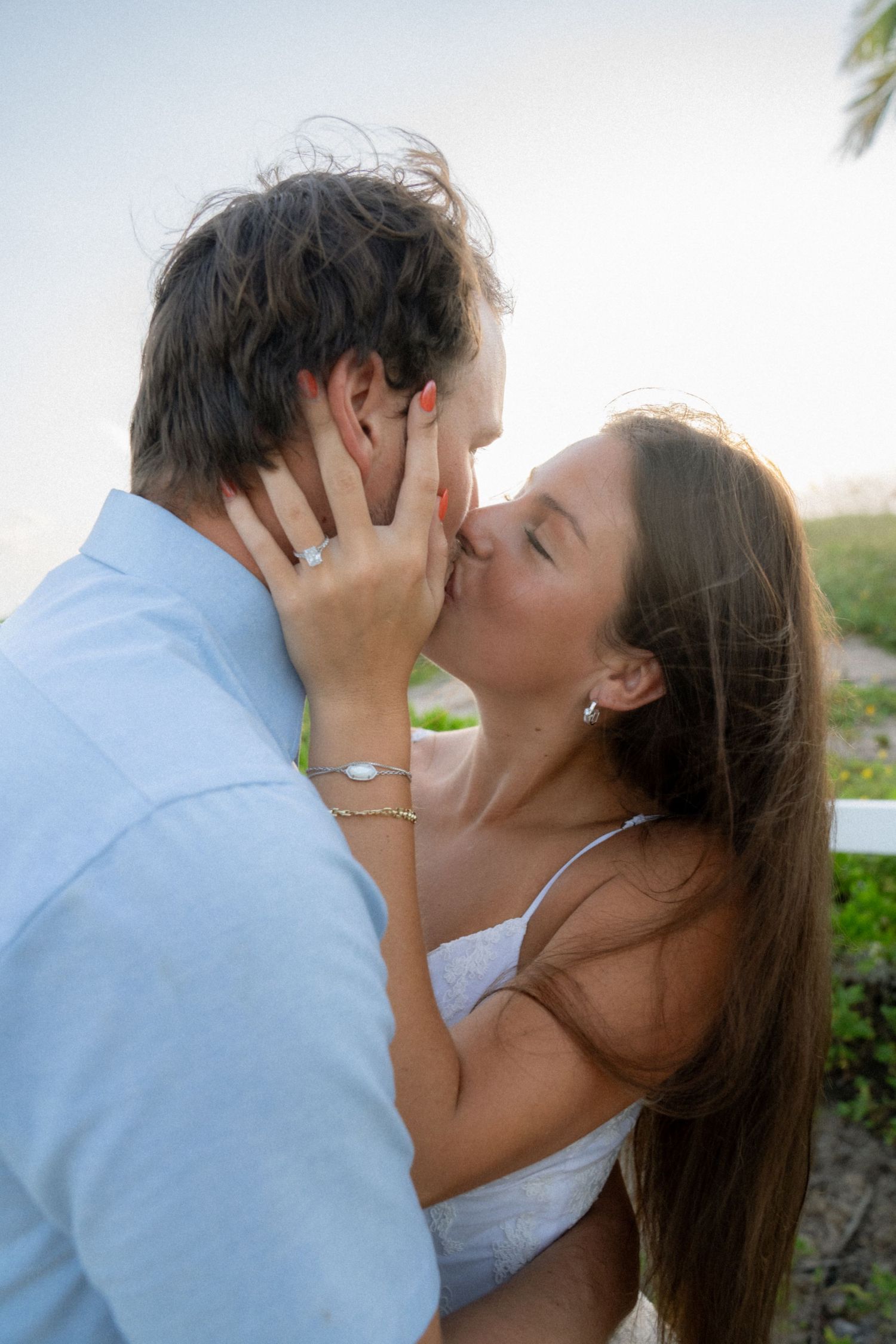 Couple sharing a romantic kiss outdoors, woman wearing engagement ring and white dress, man in light blue shirt.