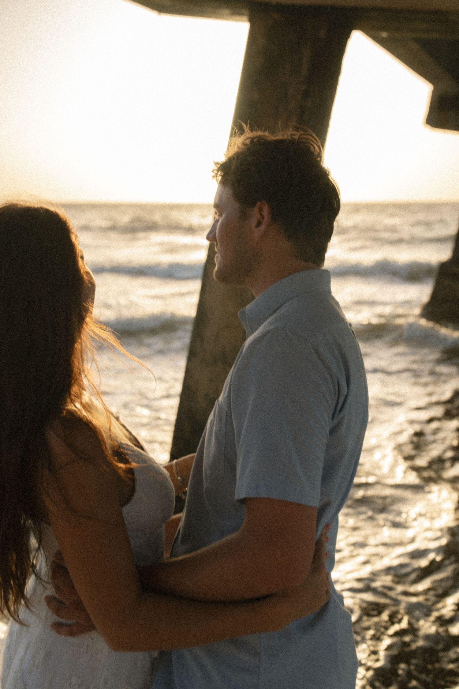 Couple embracing on beach at sunset near pier, romantic ocean backdrop with golden light.