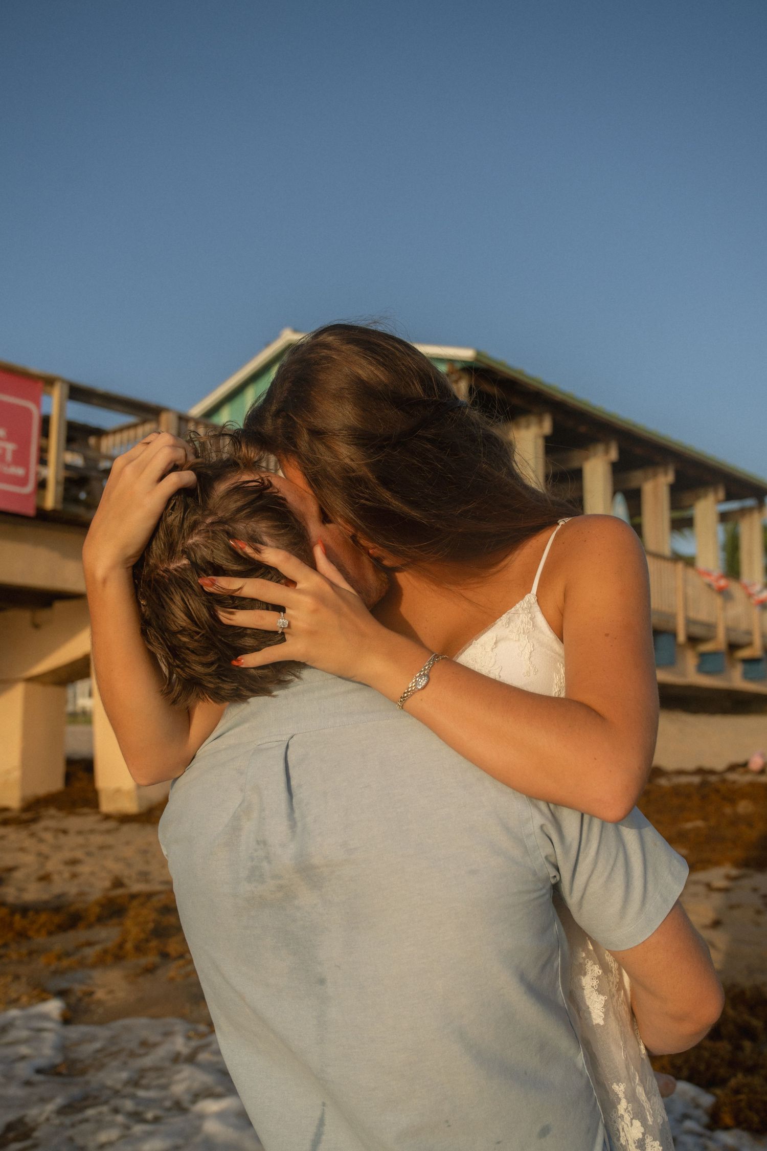 Couple sharing a romantic embrace and kiss on a beach at golden hour with a pier in the background.