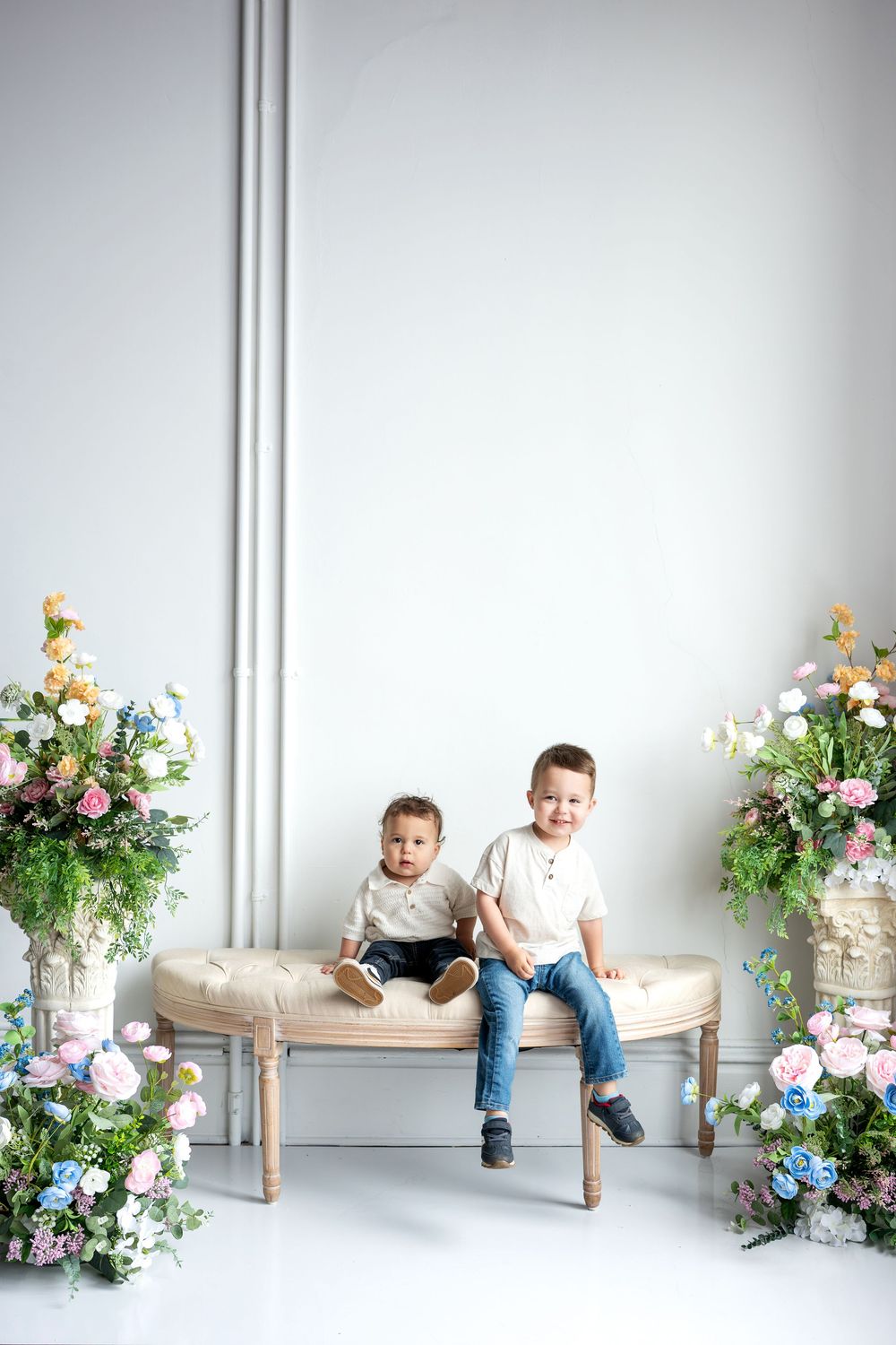 Two young brothers in white shirts and jeans sit on a bench with floral arrangements.