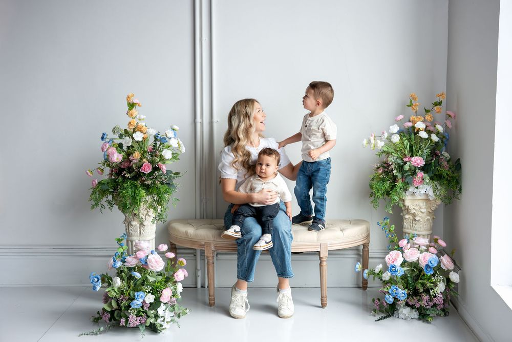 Blonde mother interacts with toddler son while holding baby on a bench with flowers.