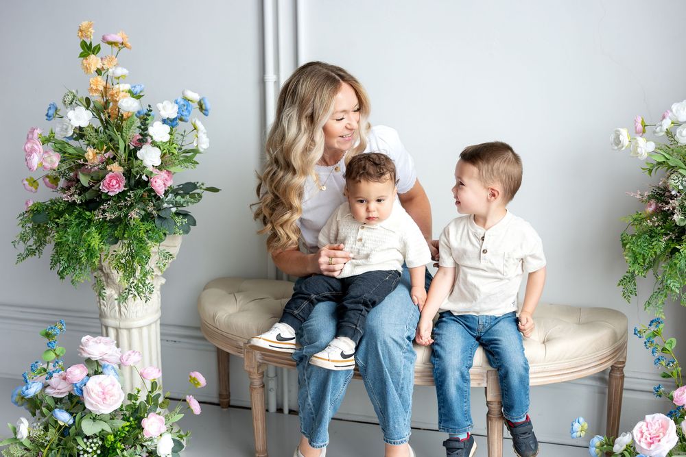Blonde mother chats with toddler son while holding baby amid colorful flower arrangements.
