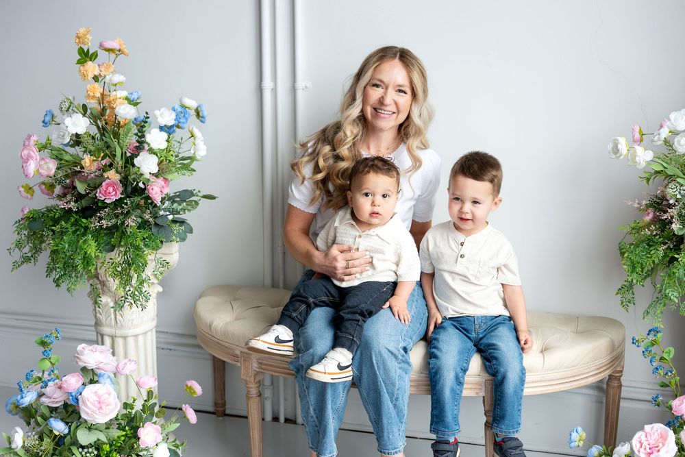 Smiling blonde mother poses with two young sons on a bench surrounded by pastel flowers.