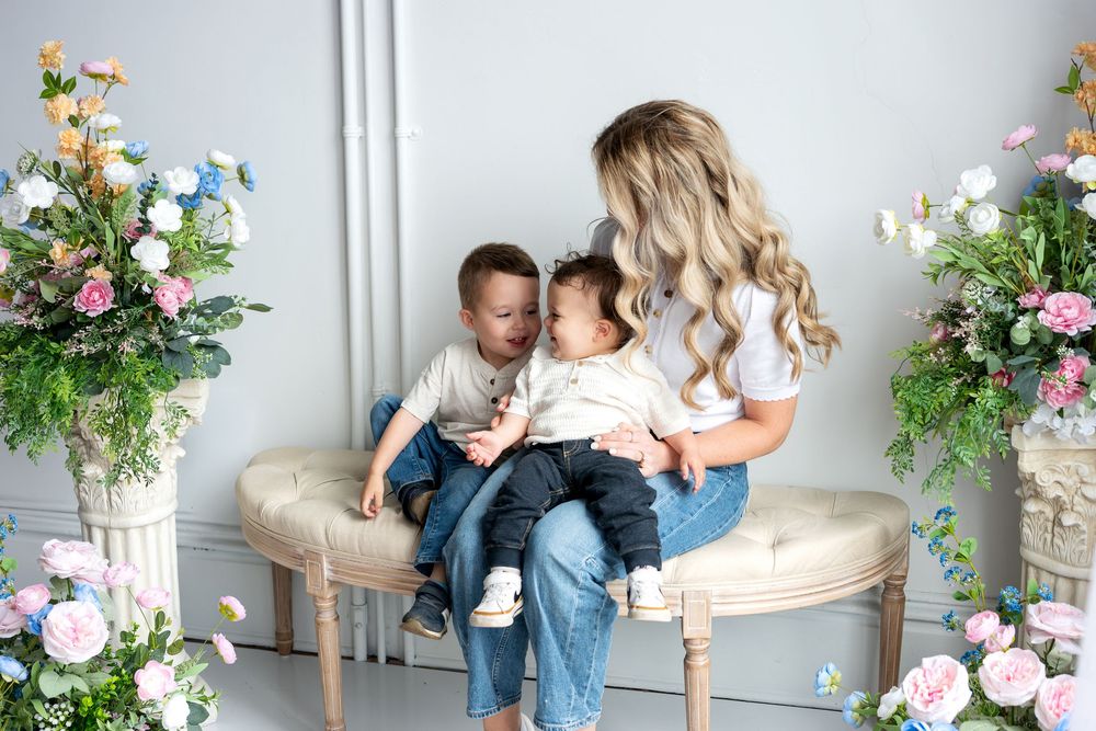 Blonde mother cuddles infant while older son leans in among elegant floral studio decor.