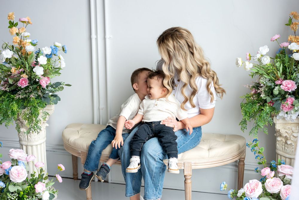A blonde mother sits on a bench kissing her two young sons surrounded by colorful floral arrangements.