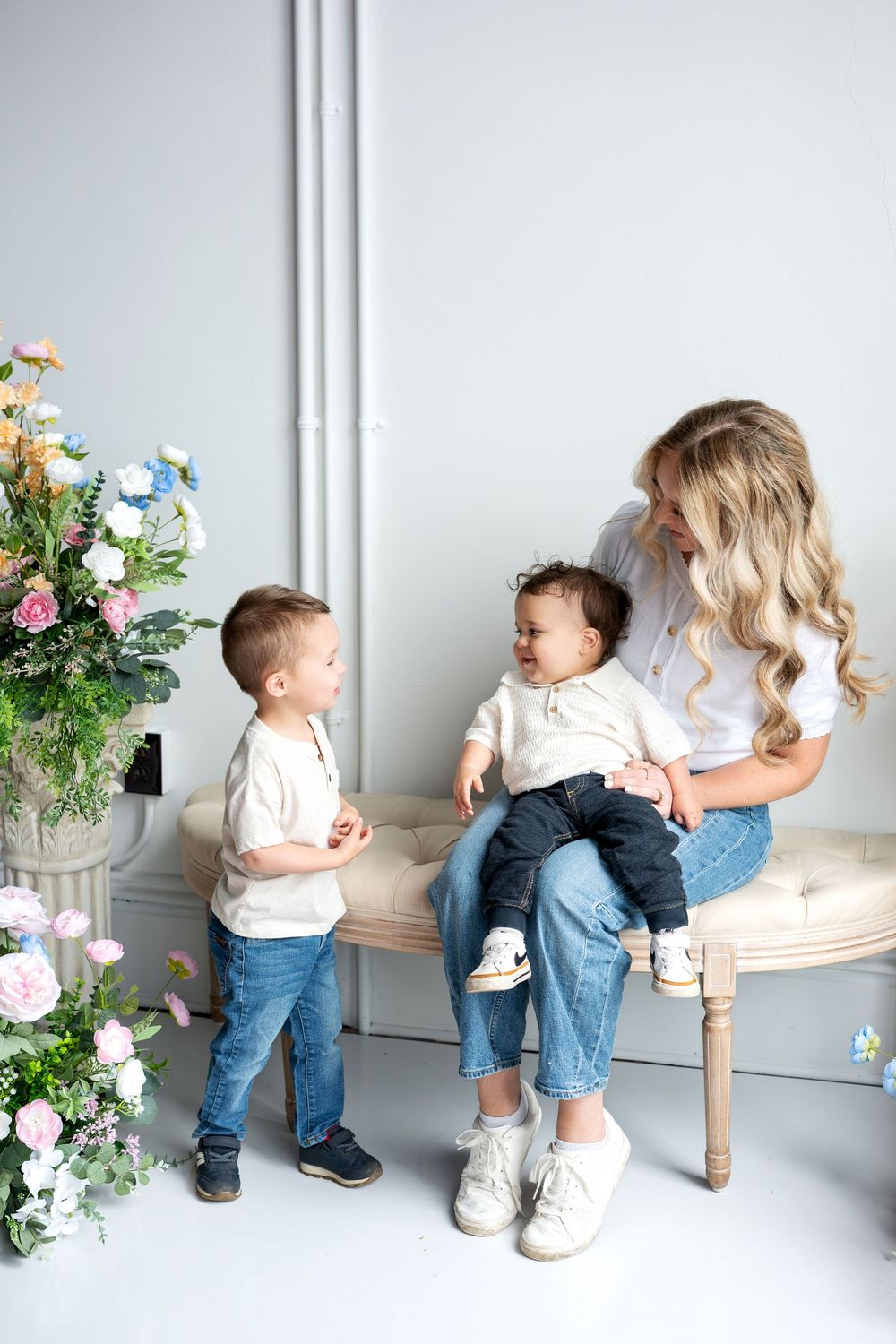 A blonde mother sits on a tufted bench with her baby while her young son stands nearby amid flowers.