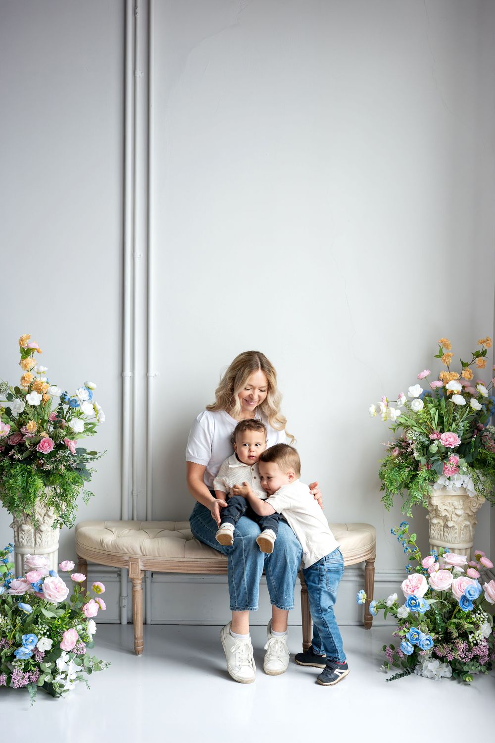 A blonde mother smiles as her toddler hugs his baby sibling on her lap on a floral studio bench.