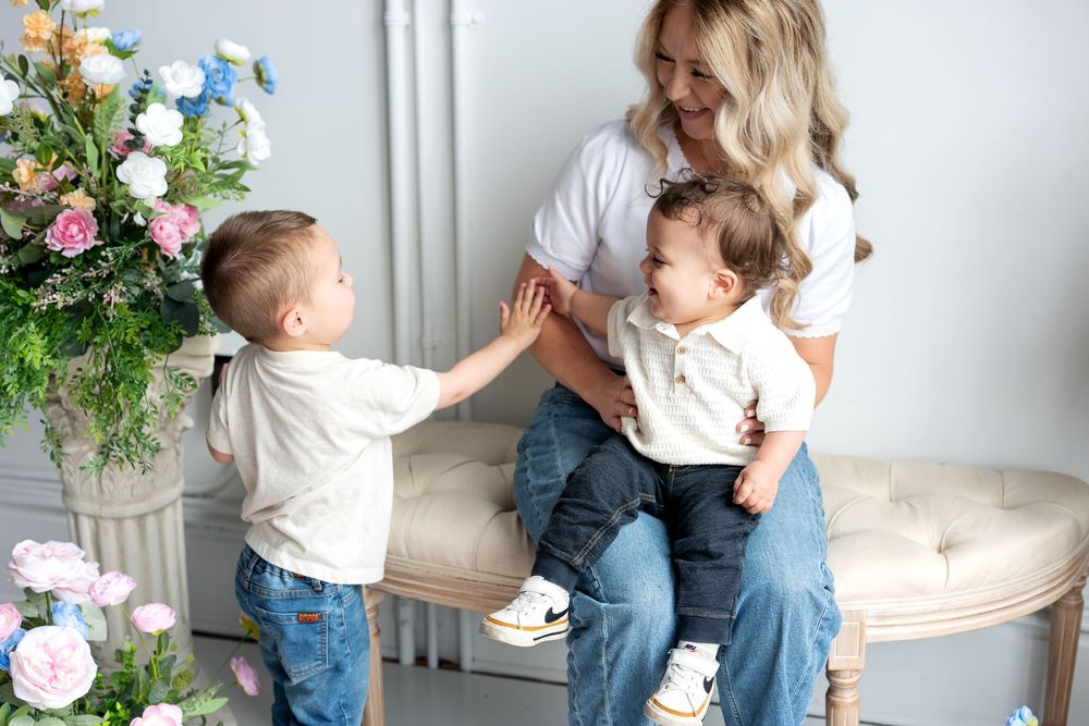 A blonde mother holds her laughing baby while her toddler son reaches out to touch the infant playfully.