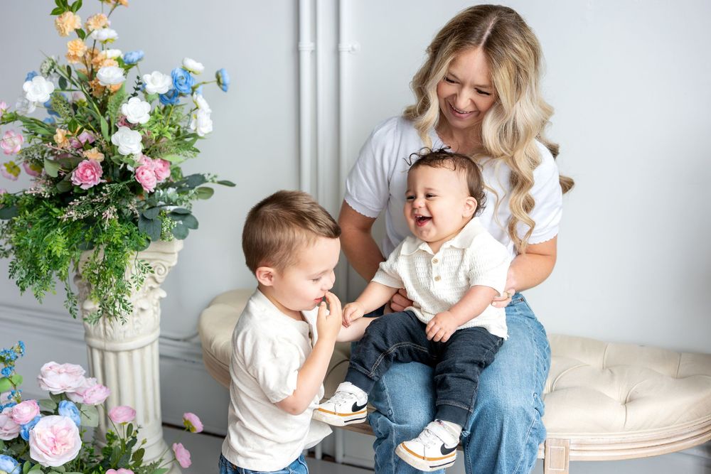 A smiling blonde mother watches her two young sons interact joyfully on a bench surrounded by flowers.