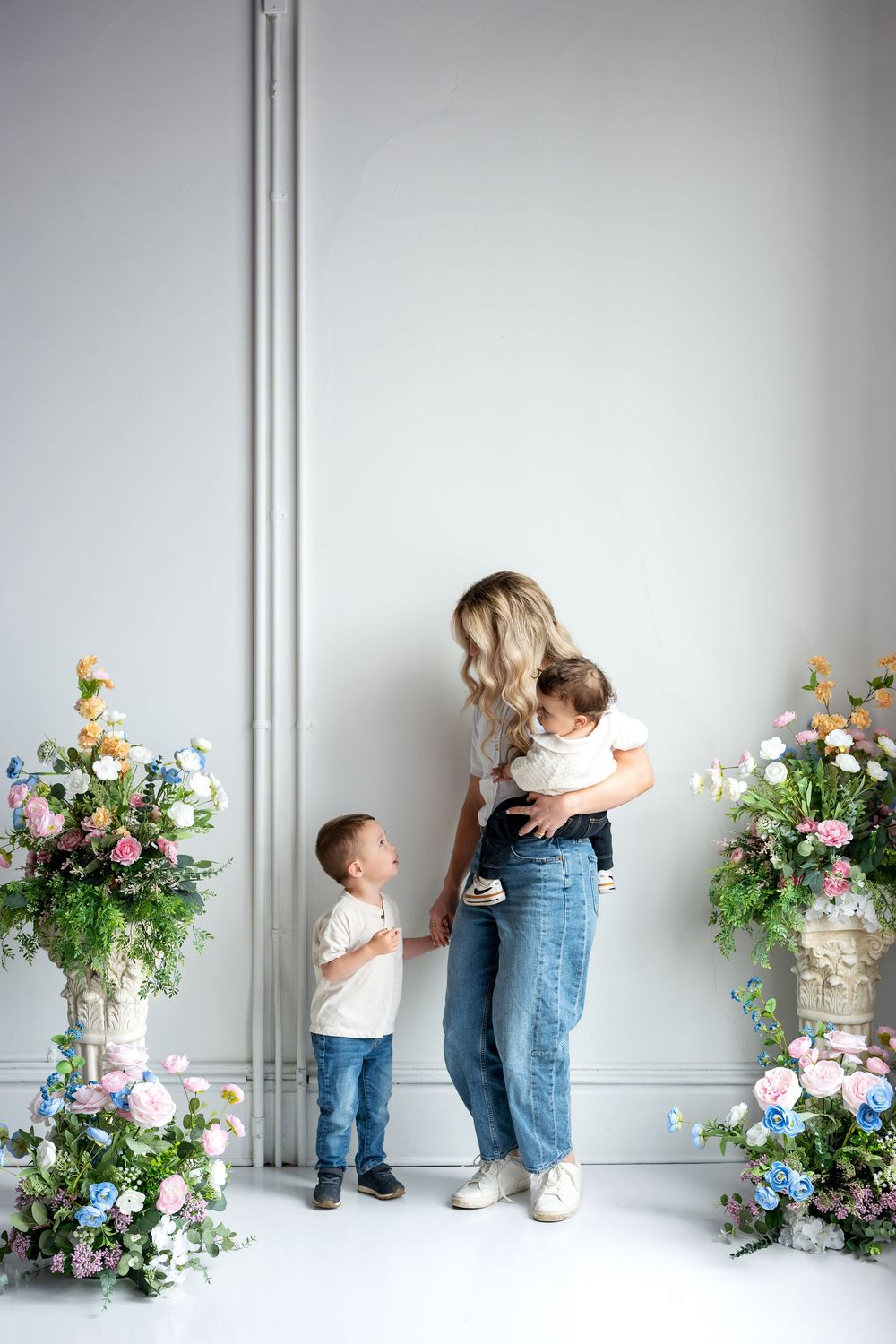 A blonde mother stands holding her baby while her young son looks up at her in a floral studio setting.