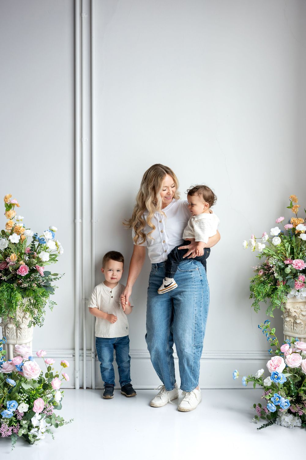 A blonde mother holds her baby and reaches for her toddler son's hand in a bright floral studio.