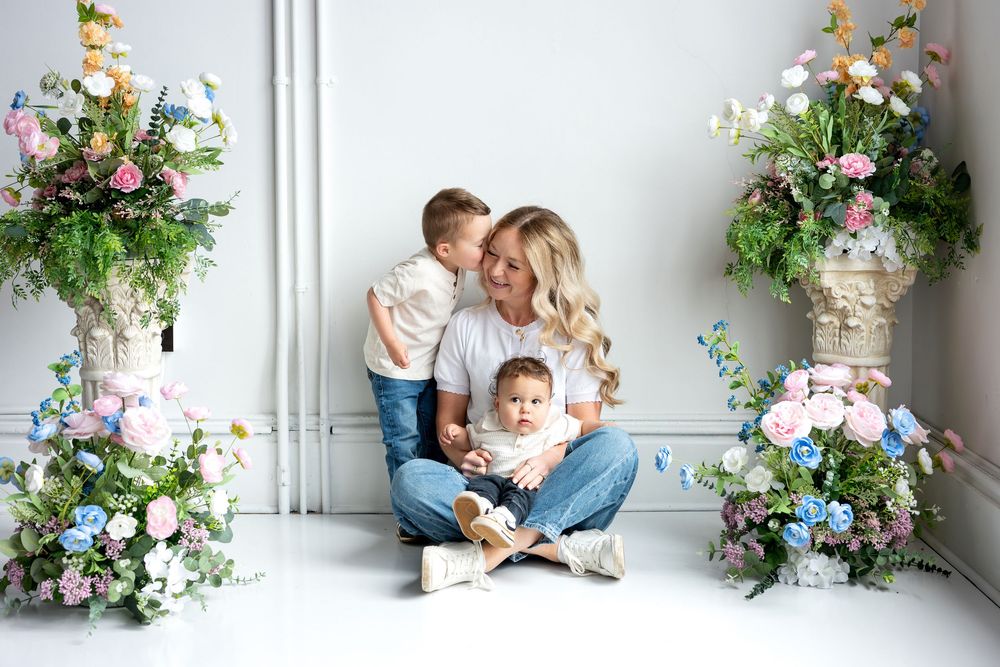 A woman with long blonde hair sits cross-legged holding two small children amid pastel flower displays in a studio.