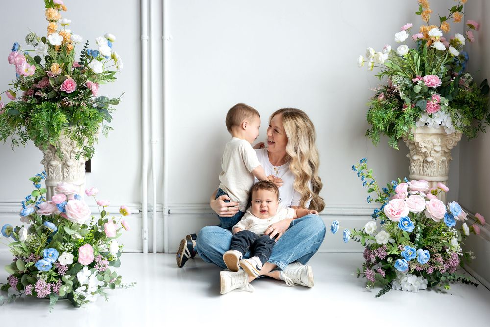 A blonde woman sits cross-legged on white floor holding two small children near tall pastel floral arrangements.