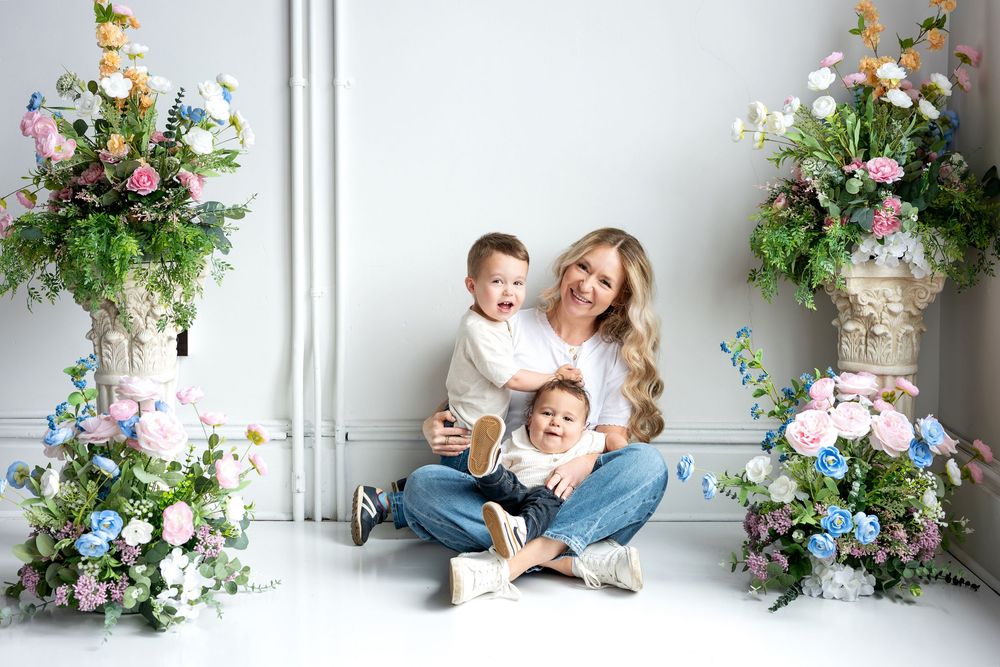 A blonde mother sits on a white floor hugging two young boys between elegant pastel floral arrangements in a studio.