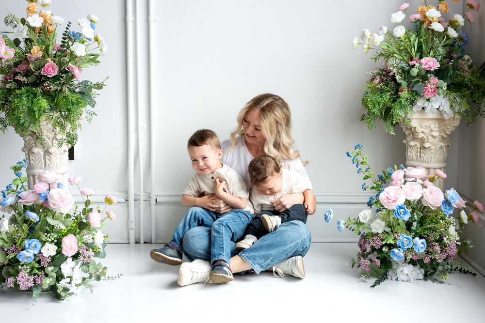 A blonde mother sits on white floor cuddling two young boys surrounded by colorful spring flower arrangements indoors.