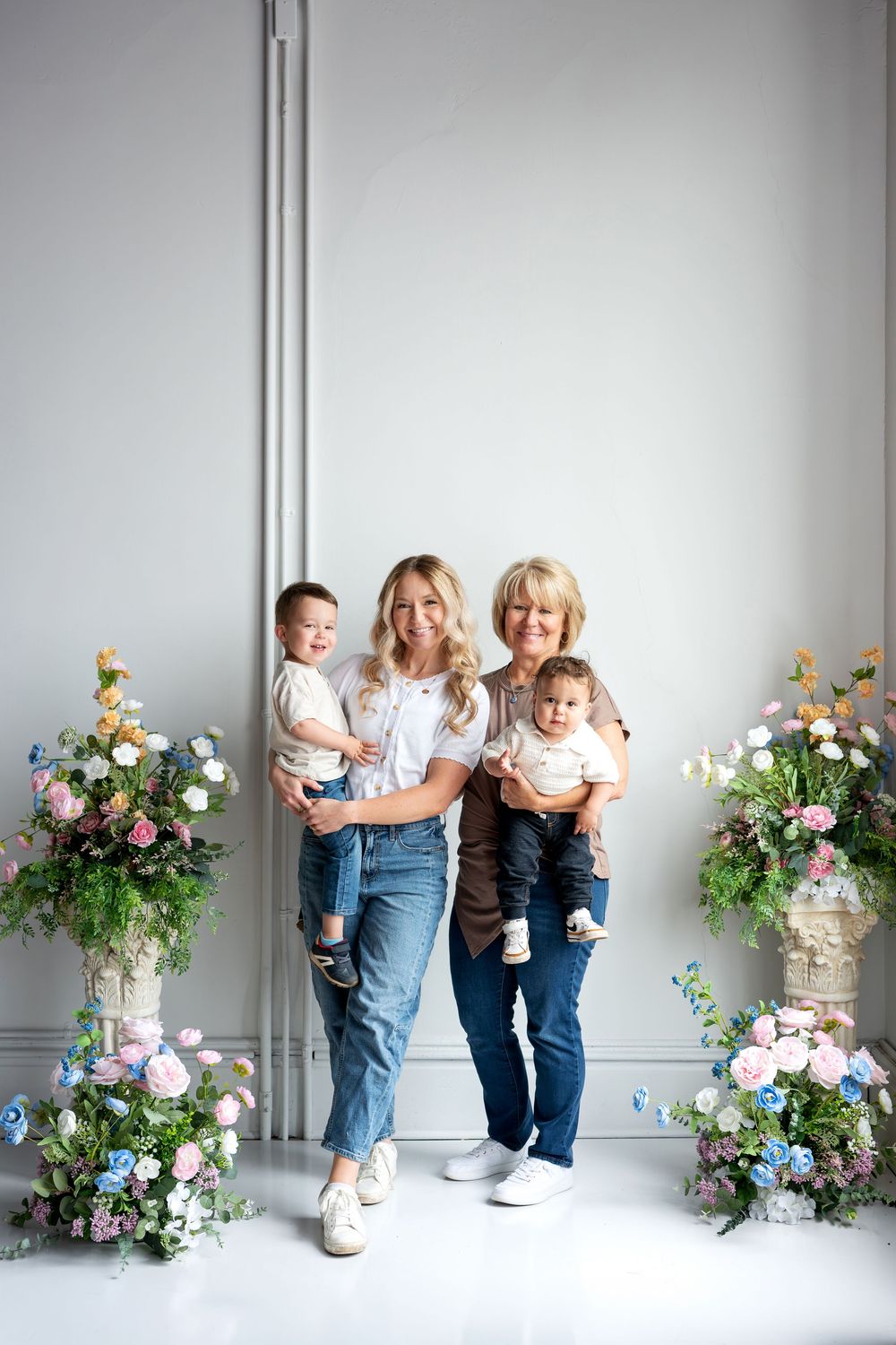 Two blonde women each hold a young child while standing between pastel floral arrangements in a bright white studio.
