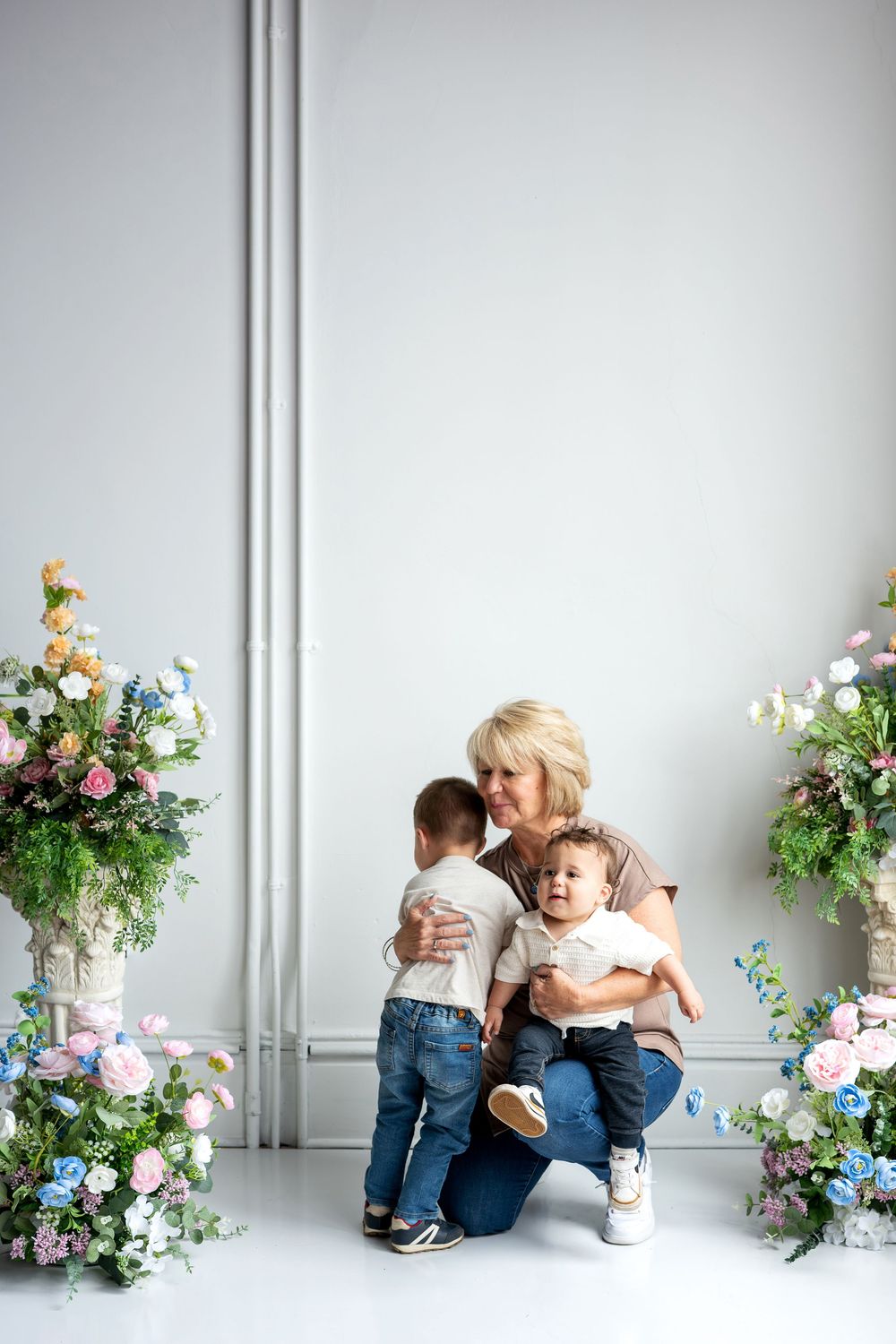 Grandmother kneeling with two toddler boys surrounded by colorful floral arrangements in a bright white studio.