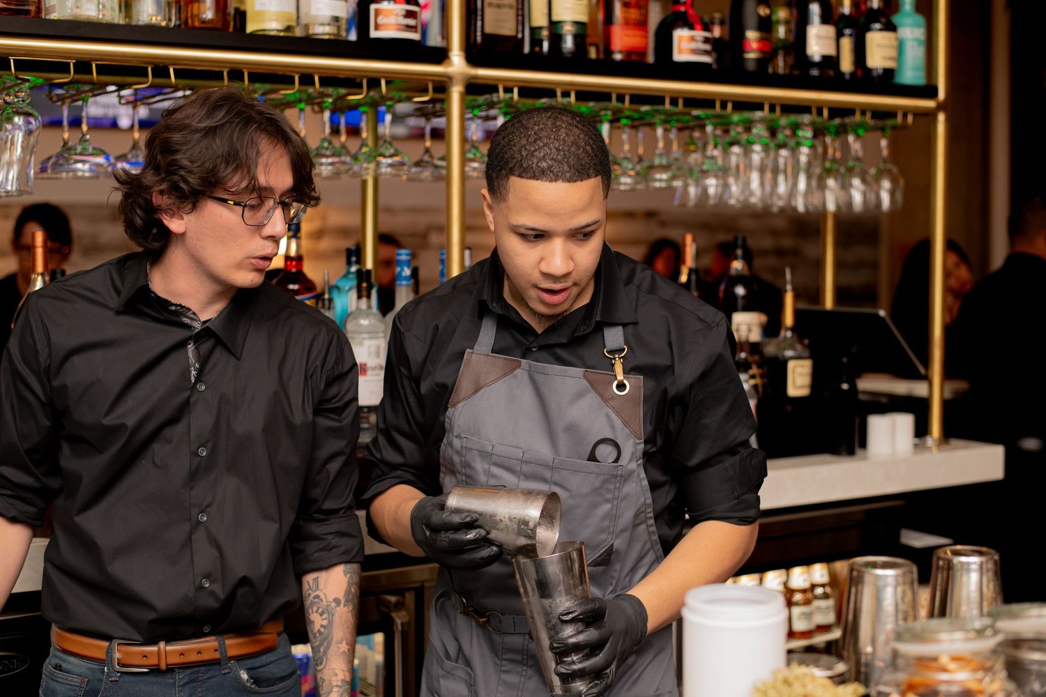 Two bartenders in black uniforms craft cocktails behind a well-stocked bar with hanging wine glasses.