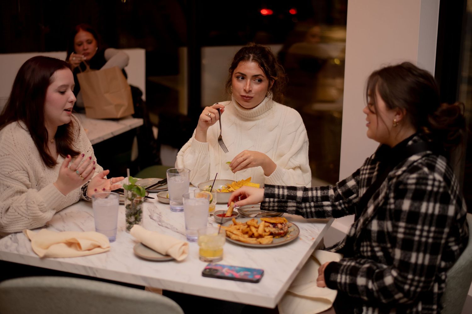 Three women dining together at a restaurant table with pizza and drinks at night.