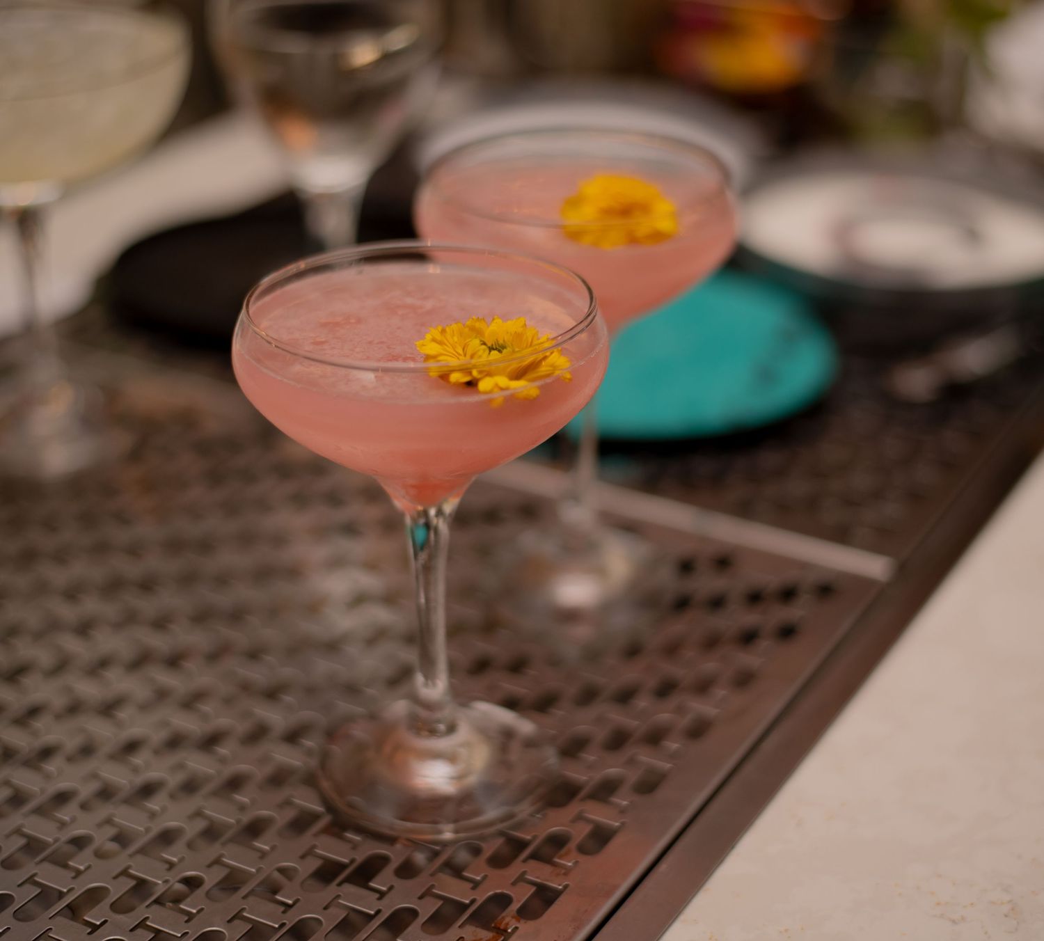 Two elegant pink cocktails garnished with yellow flowers served in coupe glasses.