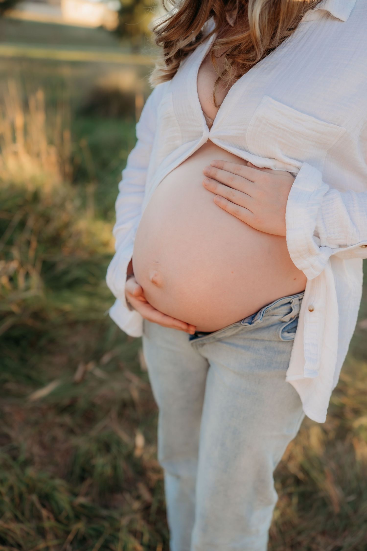 Pregnant woman in white shirt and jeans cradling bare baby bump outdoors in golden sunlight.