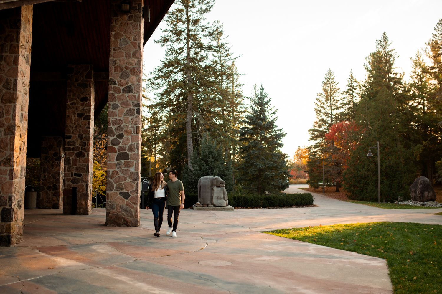 Two people walking along a stone building pathway surrounded by tall evergreen trees in a scenic autumn campus setting.