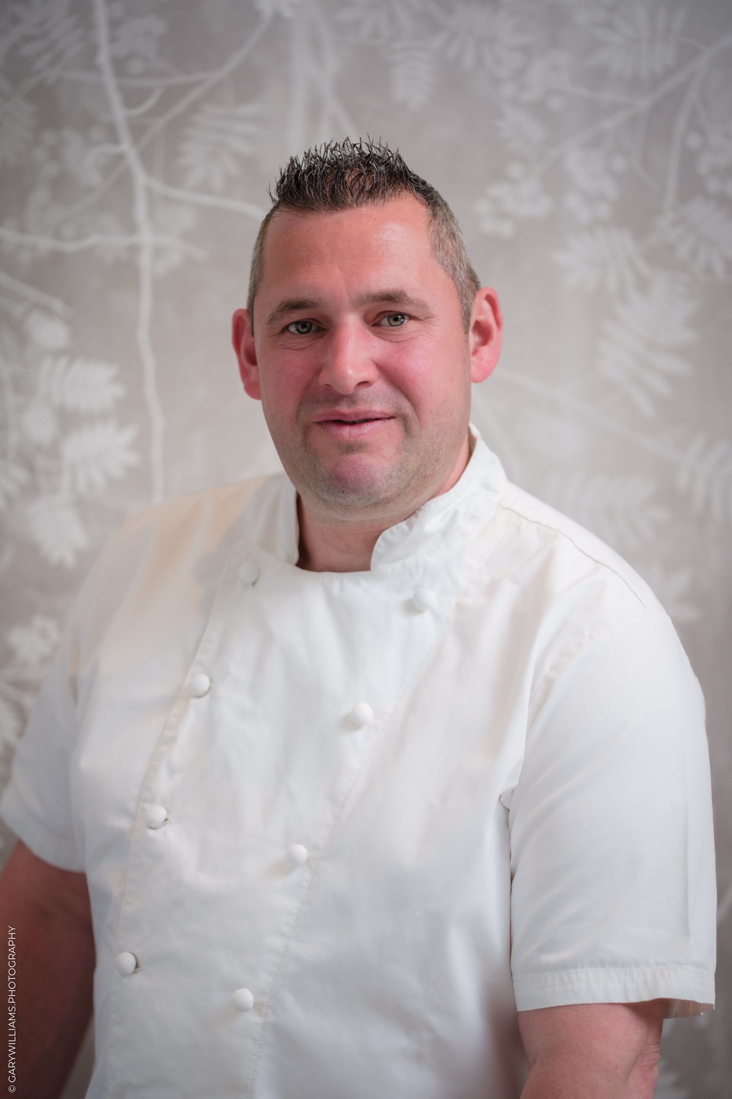 A professional chef in a white double-breasted chef coat smiles confidently against a decorative floral background.