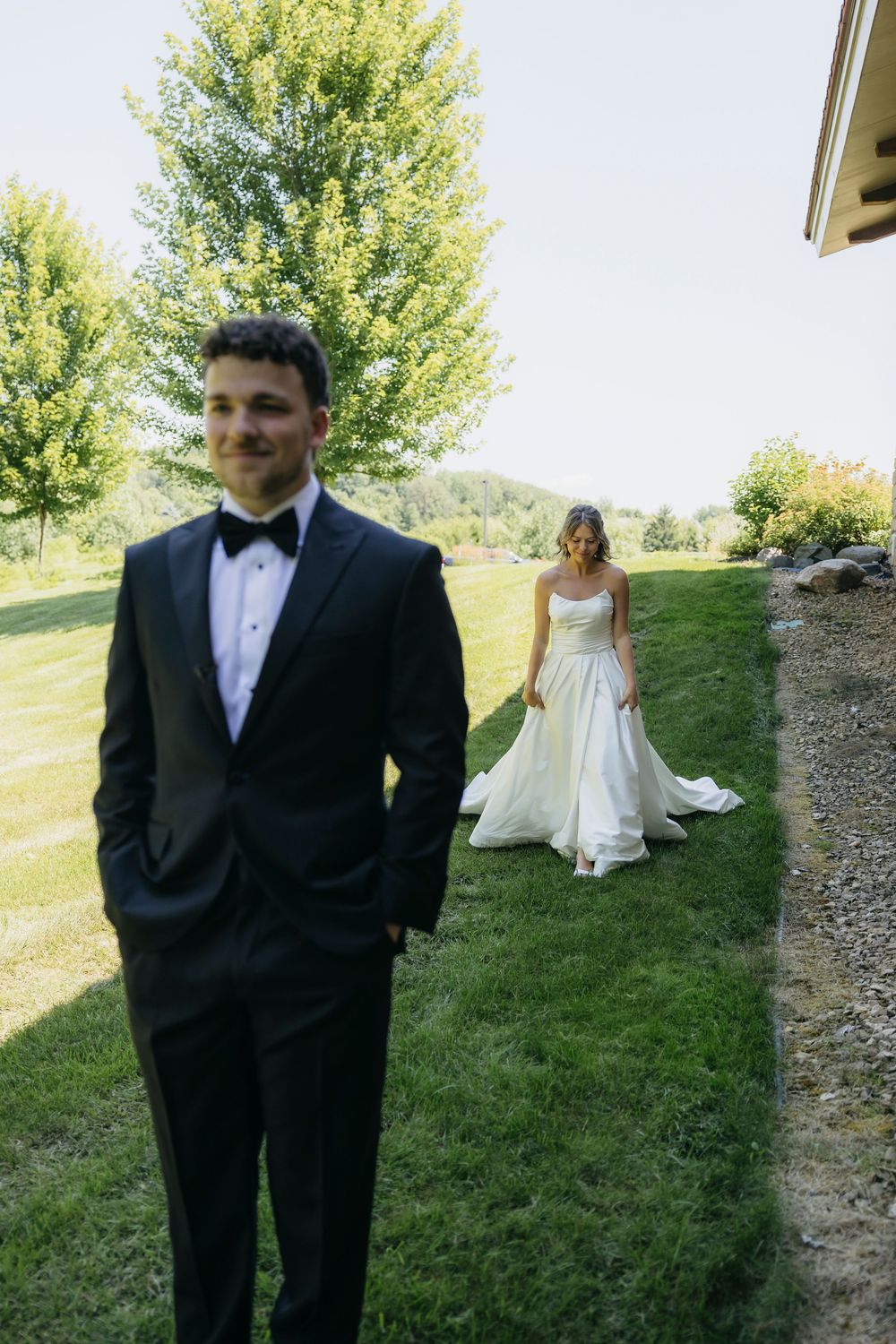 Groom in black tuxedo stands outdoors as bride in white wedding gown approaches on green lawn before their first look
