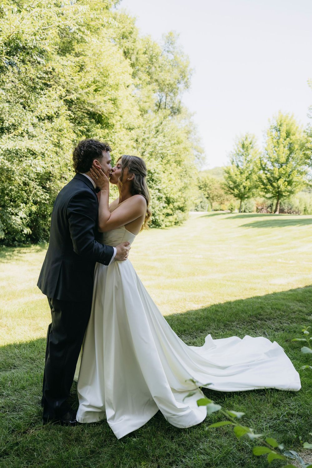 Bride and groom share a romantic kiss outdoors on lush green grass, surrounded by trees on their wedding day after the first look