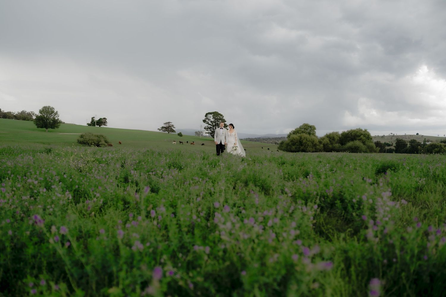 Couple in wedding attire walking through a lush purple wildflower field under dramatic storm clouds in the countryside.