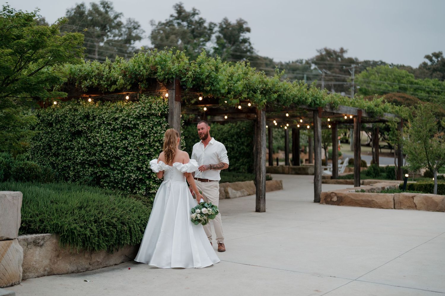 Bride in white gown and groom in tan suit exchange vows under string lights in a lush garden venue.